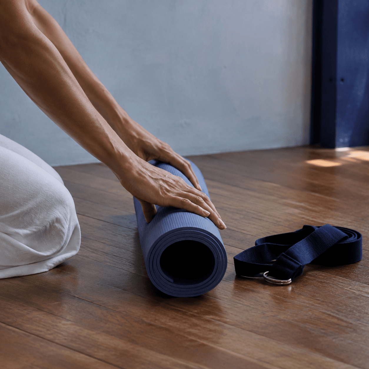 Hands rolling an indigo yoga mat beside a neatly placed practice strap